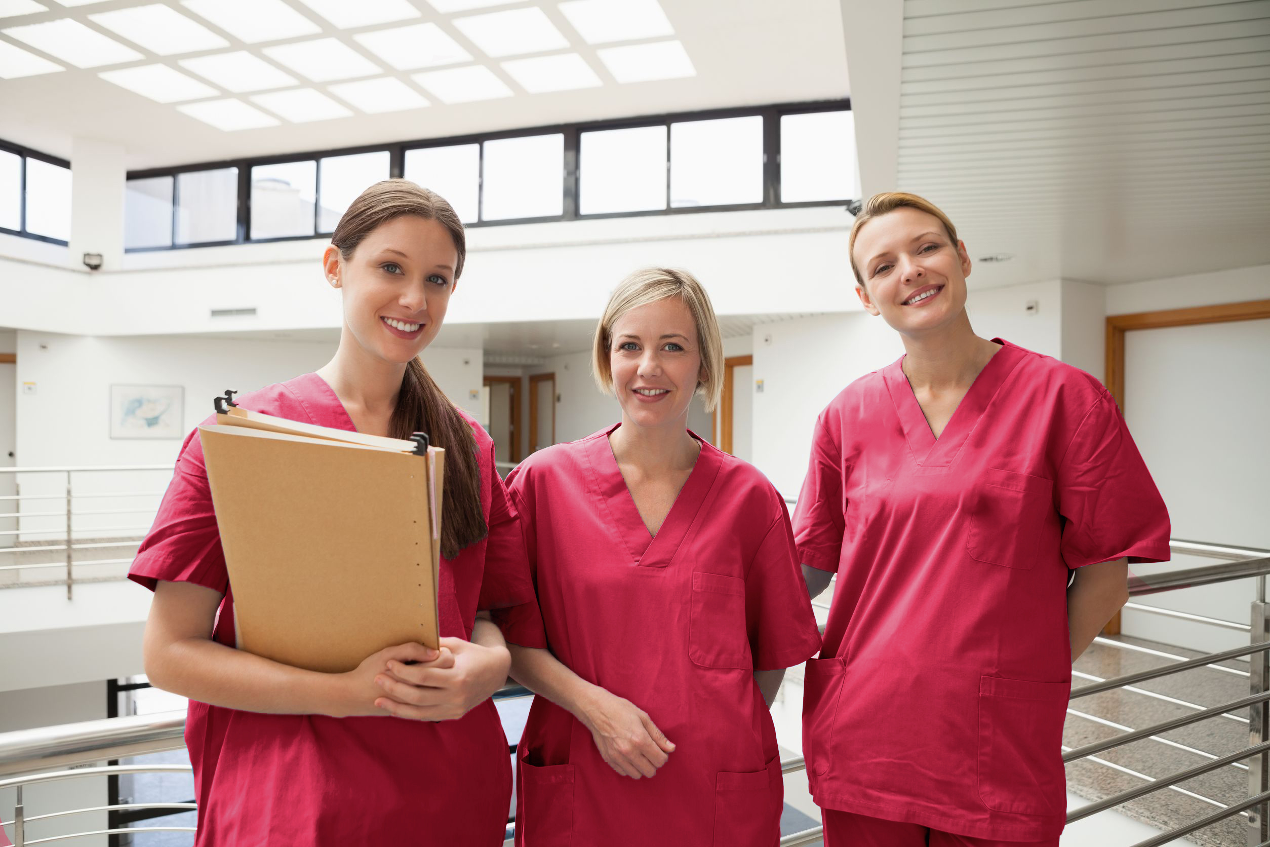 Drei Pflegekräfte in roter Arbeitskleidung stehen lächelnd in einem hellen, modernen Gebäude. Eine der Frauen hält eine Akte in der Hand. Die Atmosphäre wirkt freundlich und professionell.