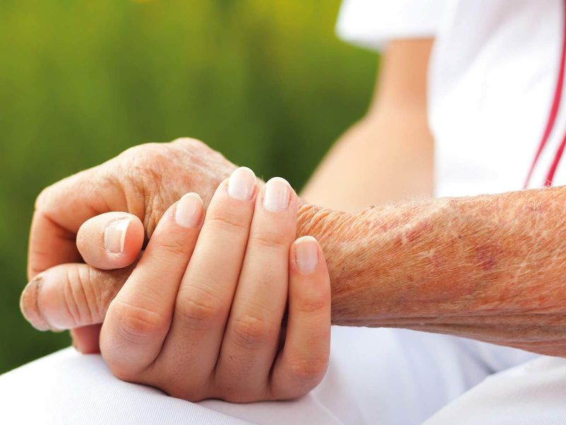 Eine jüngere Person hält fürsorglich die Hand einer älteren Person. Die Szene vermittelt Nähe, Unterstützung und Mitgefühl. Der Hintergrund ist unscharf und grün, was auf eine ruhige Umgebung im Freien hindeutet.