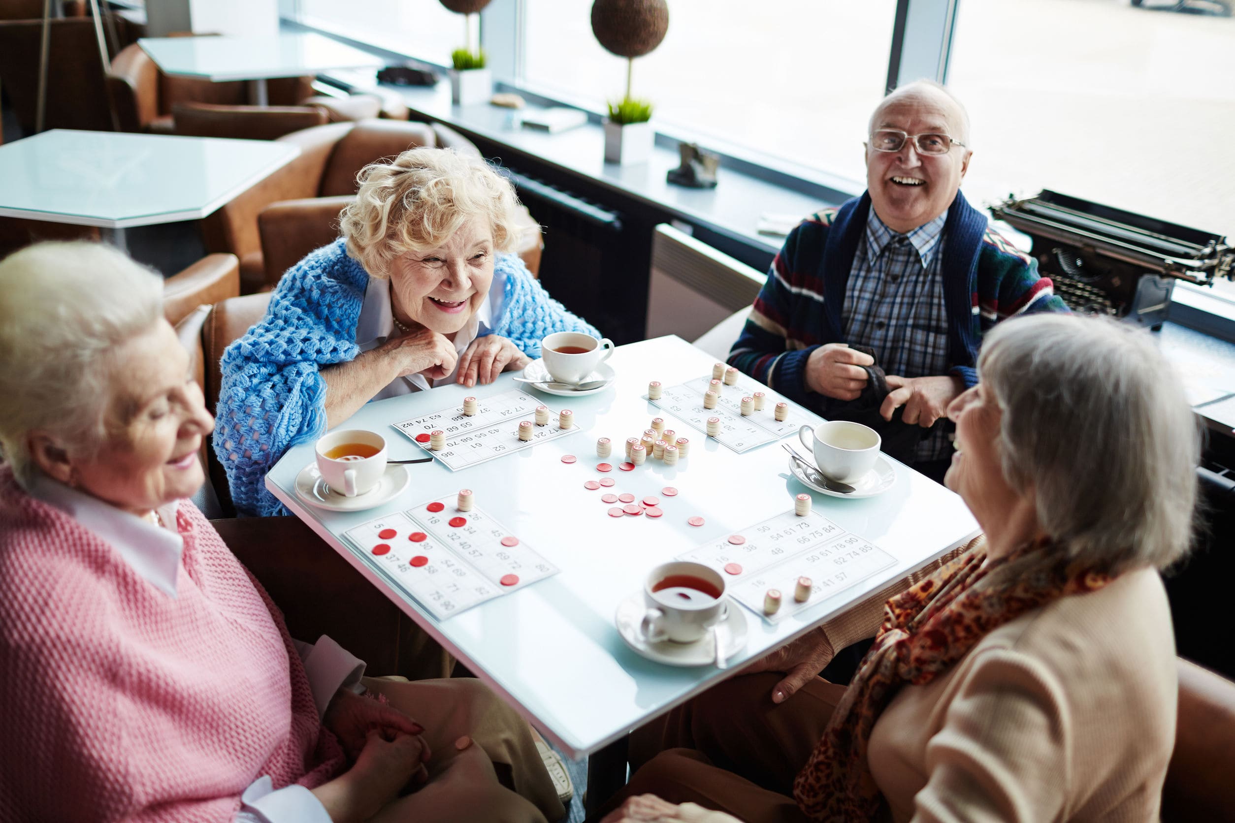 Vier ältere Menschen sitzen gemeinsam an einem Tisch in einem Café oder Aufenthaltsraum. Sie unterhalten sich fröhlich, trinken Tee oder Kaffee und spielen ein Gesellschaftsspiel. Die Stimmung ist gesellig und heiter.