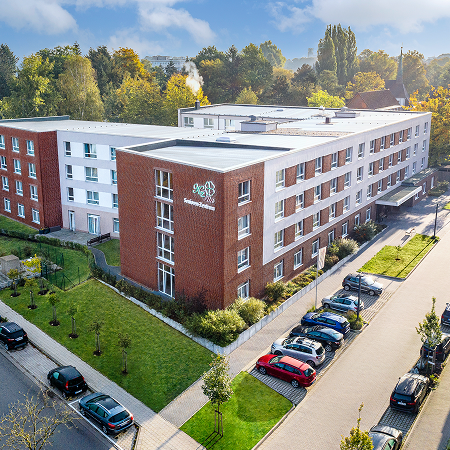 Luftaufnahme eines modernen, mehrstöckigen Gebäudes mit roter und weißer Fassade. Vor dem Haus stehen mehrere Autos, und das Gelände ist von gepflegtem Grün und Bäumen umgeben. Es wirkt wie eine Pflegeeinrichtung oder ein Seniorenwohnheim.
