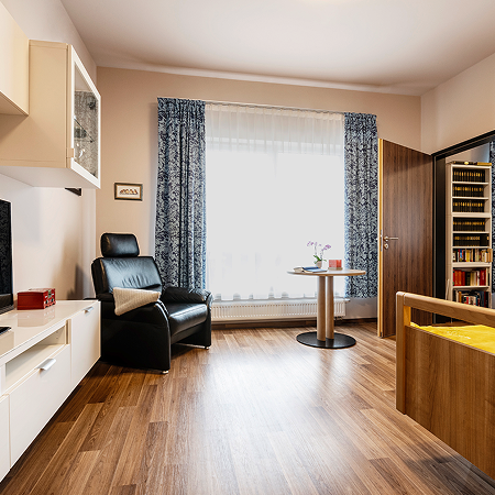 Gemütlich eingerichtetes Zimmer mit Holzfußboden, einem Pflegebett, einem schwarzen Fernsehsessel, einem kleinen Tisch mit zwei Stühlen und einem großen Fenster mit blauen Vorhängen. Die Atmosphäre wirkt hell, wohnlich und einladend.