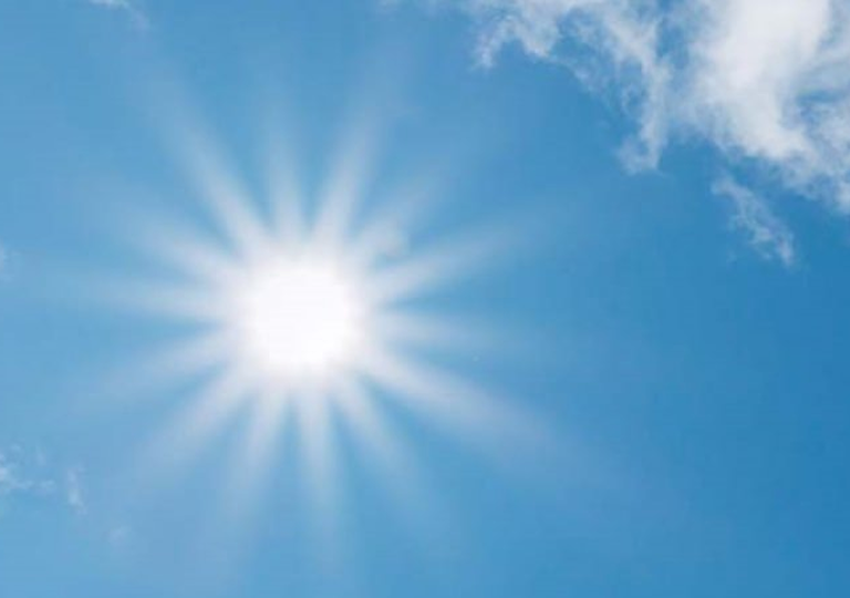 Strahlend blauer Himmel mit wenigen weißen Wolken und einer hell leuchtenden Sonne, die Lichtstrahlen in alle Richtungen aussendet – eine Szene, die für schönes Wetter und Sommerstimmung steht.