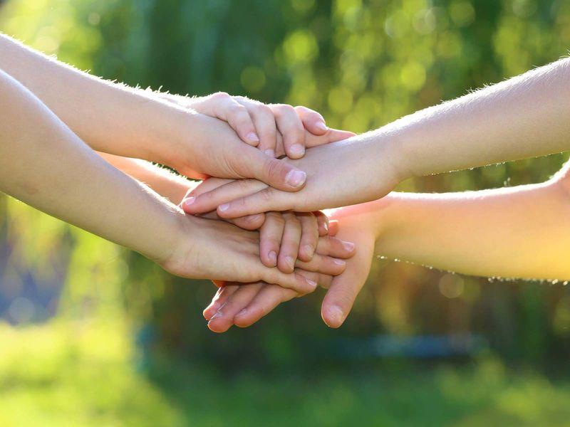 Mehrere Hände von Kindern oder Jugendlichen liegen übereinander in einer Geste des Zusammenhalts. Der Hintergrund ist grün und sonnig, was eine freundliche und naturverbundene Atmosphäre vermittelt.