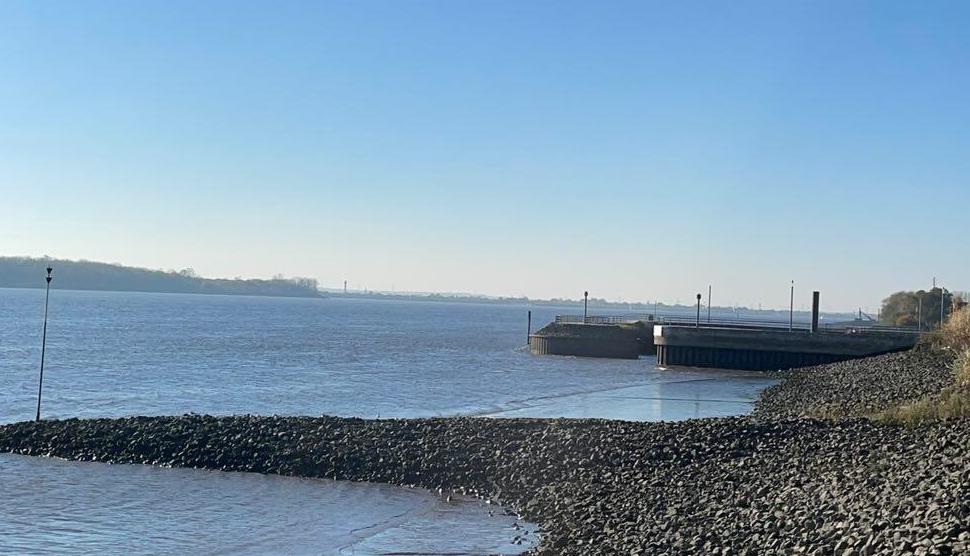 Blick auf einen ruhigen Küstenabschnitt mit Kieselstrand und einem kleinen Pier, der ins Wasser ragt. Im Hintergrund sind sanfte Hügel und das weite Meer unter klarem, blauem Himmel zu sehen – eine friedliche und klare Küstenlandschaft.