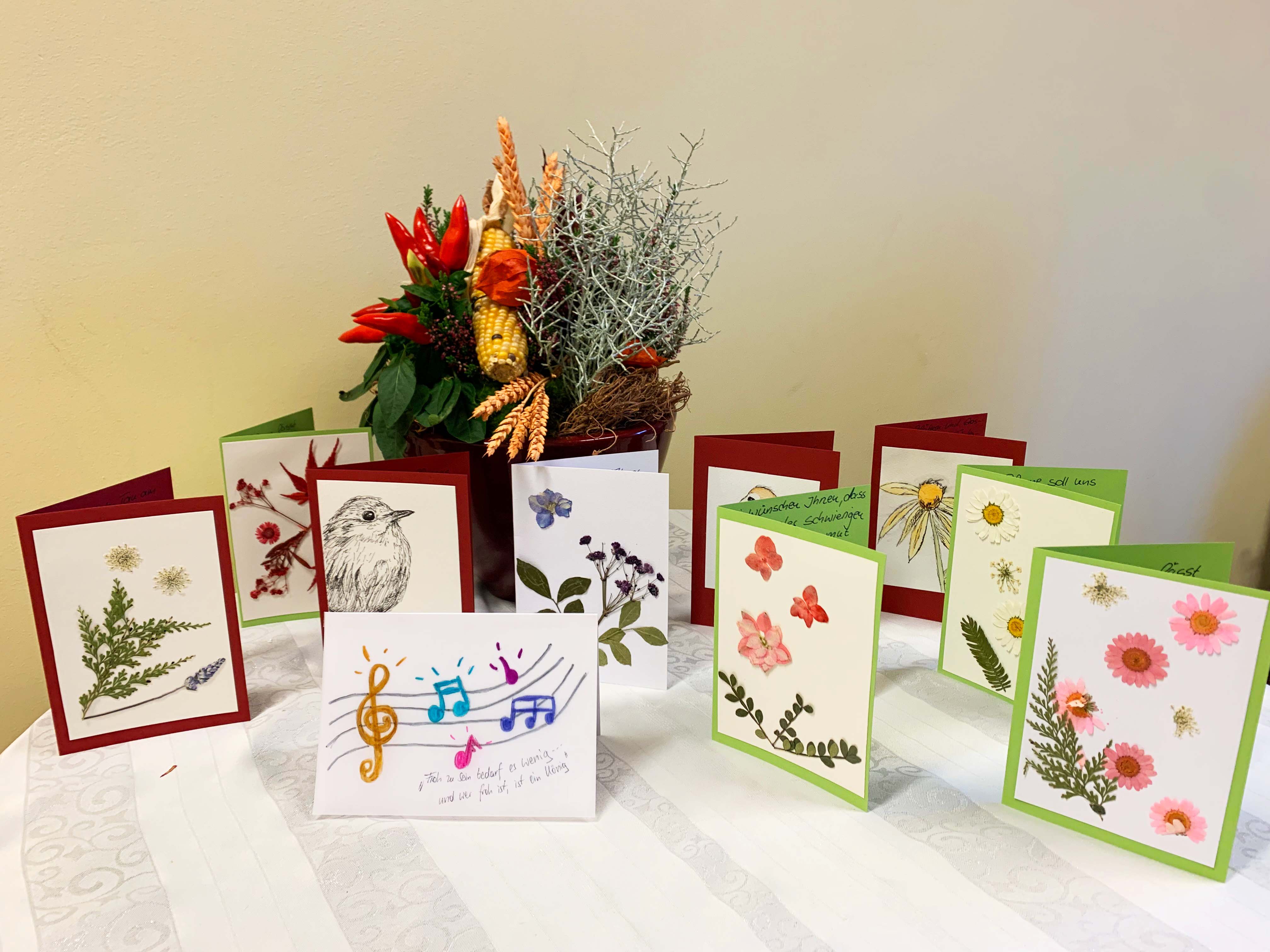 Tisch mit selbstgebastelten, bunten Grußkarten, die mit Blumen- und Naturmotiven verziert sind. Im Hintergrund steht eine herbstliche Blumendekoration mit Zweigen und roten Blüten. Die Szene vermittelt Kreativität und saisonale Gestaltung.