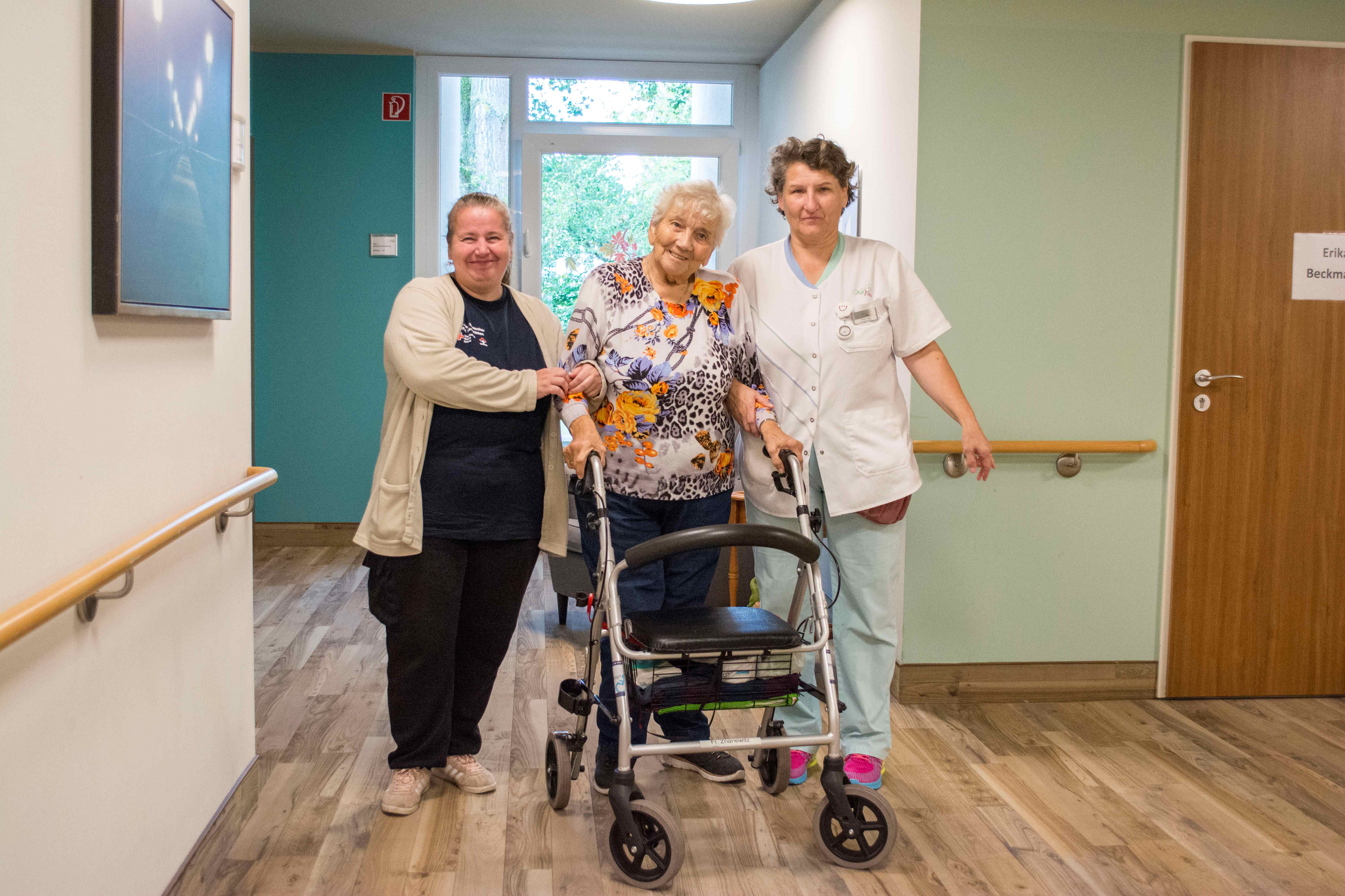 Eine ältere Frau mit Rollator steht lächelnd zwischen zwei Betreuerinnen in einem hellen Flur einer Pflegeeinrichtung. Sie hält einen bunten Blumenstrauß – die Szene vermittelt Freude, Unterstützung und Gemeinschaft.