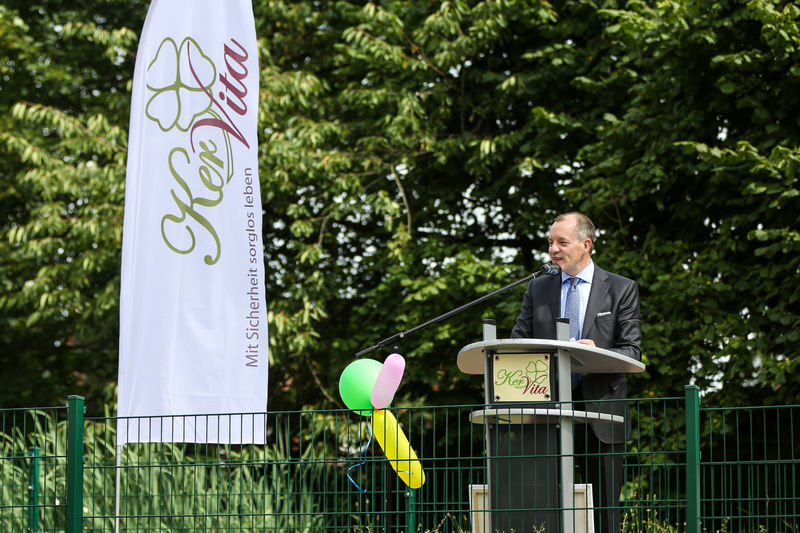 Ein Mann im Anzug steht an einem Rednerpult im Freien und hält eine Rede. Links daneben weht eine Fahne mit dem Logo der „KerVita Gruppe“. Im Hintergrund sind grüne Bäume zu sehen – die Szene wirkt feierlich und offiziell.
