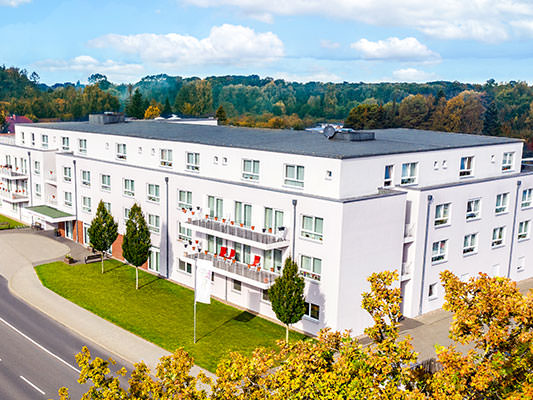 Moderne, weiß verputzte Pflegeeinrichtung mit mehreren Stockwerken an einer Straße gelegen. Der gepflegte Vorgarten ist mit Bäumen und Rasenflächen gestaltet. Im Hintergrund sind Hügel und herbstlich gefärbte Wälder zu sehen – die Szene wirkt ruhig und fr