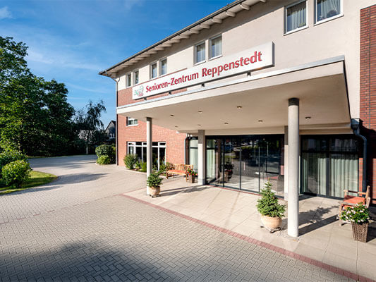 Eingangsbereich eines modernen Pflegeheims mit überdachtem Vordach und der Beschriftung „Haus am Park – Senioren-Zentrum“ über dem Eingang. Der Bereich ist mit Blumen und Sitzgelegenheiten gestaltet, die Umgebung ist grün und gepflegt.