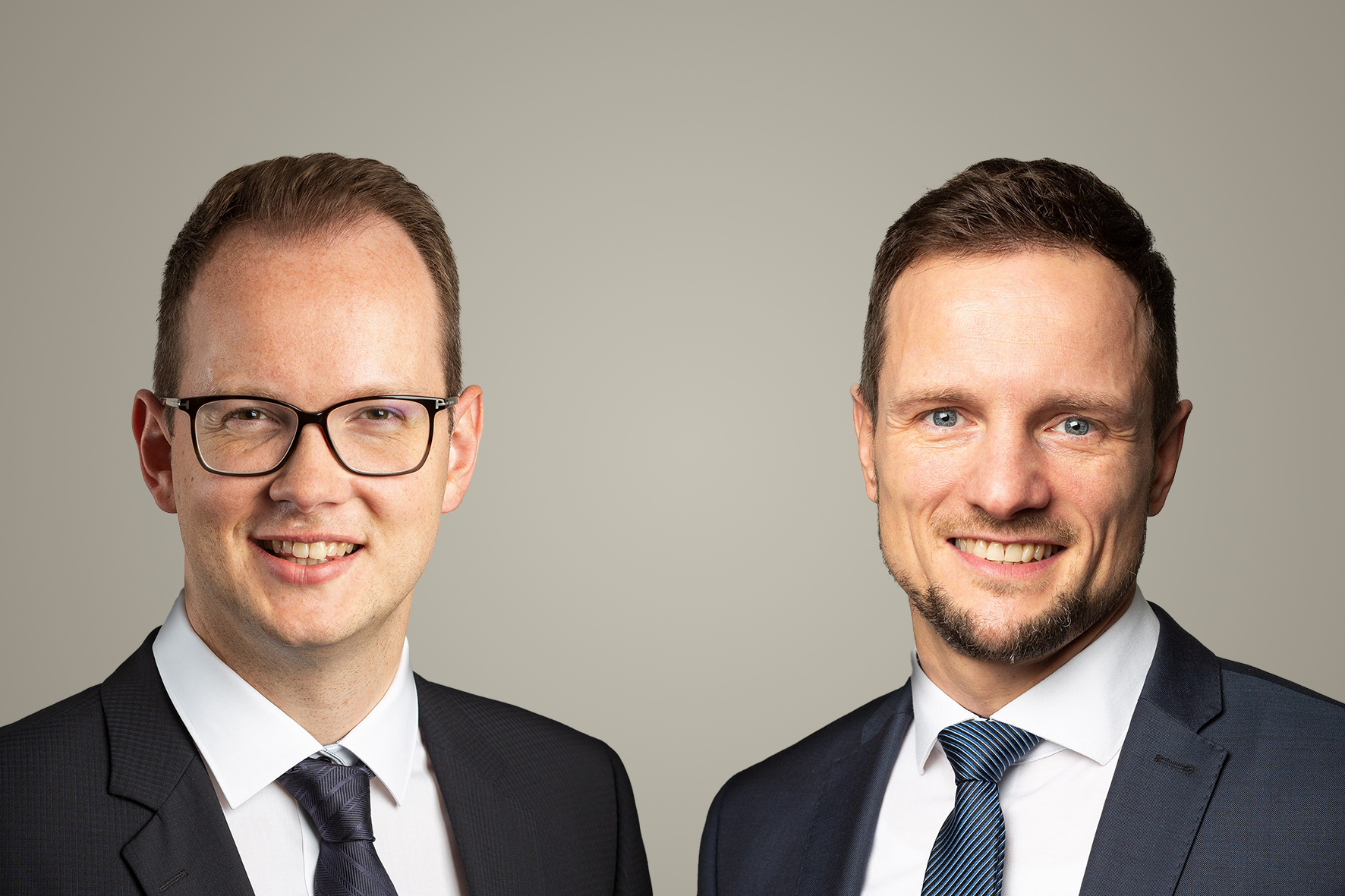 Zwei Männer in dunklen Anzügen und Krawatten posieren lächelnd vor einem neutralen, hellgrauen Hintergrund. Der linke Mann trägt eine Brille, beide wirken professionell und freundlich – ein klassisches Business-Porträt.
