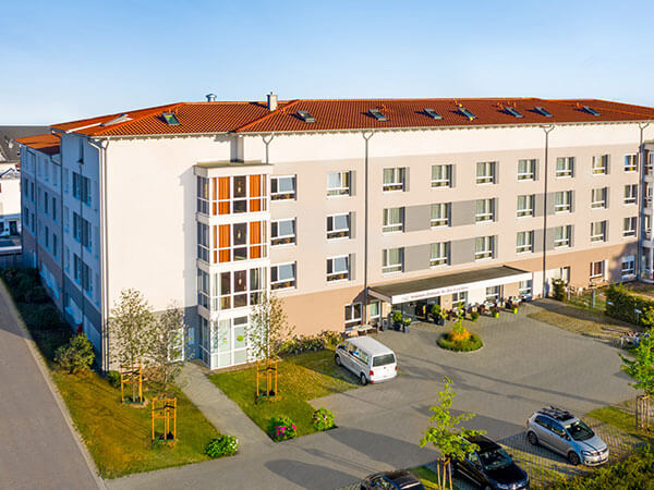 Luftaufnahme eines modernen, vierstöckigen Gebäudes mit rotem Satteldach, hellen Fassaden und einem Eingangsbereich mit Vordach. Vor dem Gebäude befinden sich Parkplätze und gepflegte Grünflächen – die Szene zeigt eine Senioreneinrichtung in ruhiger Umge