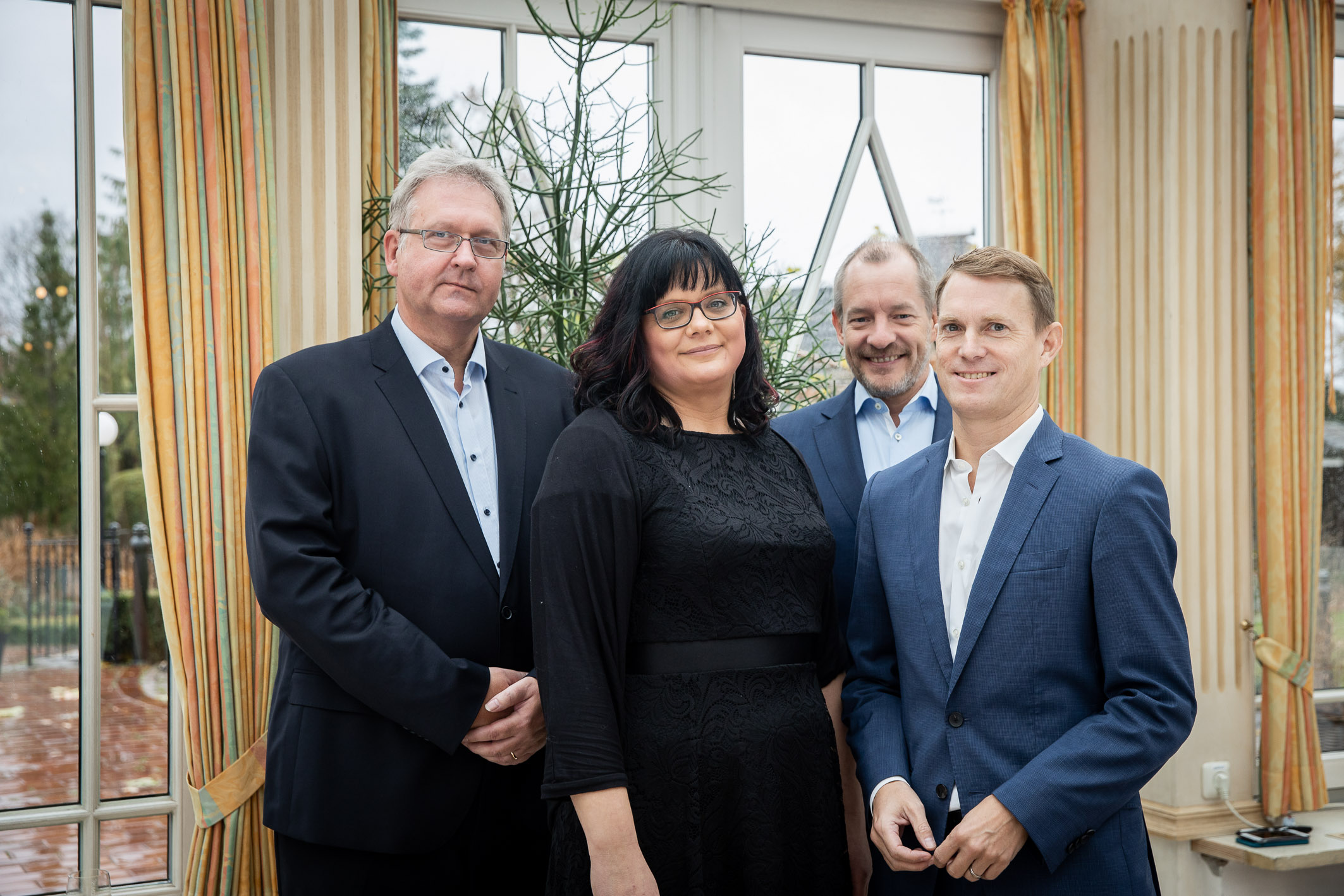 Vier Personen in eleganter Kleidung stehen gemeinsam in einem hellen Raum mit großen Fenstern und goldfarbenen Vorhängen. Sie blicken freundlich in die Kamera – die Szene wirkt professionell und zugleich herzlich, möglicherweise bei einer geschäftlichen o