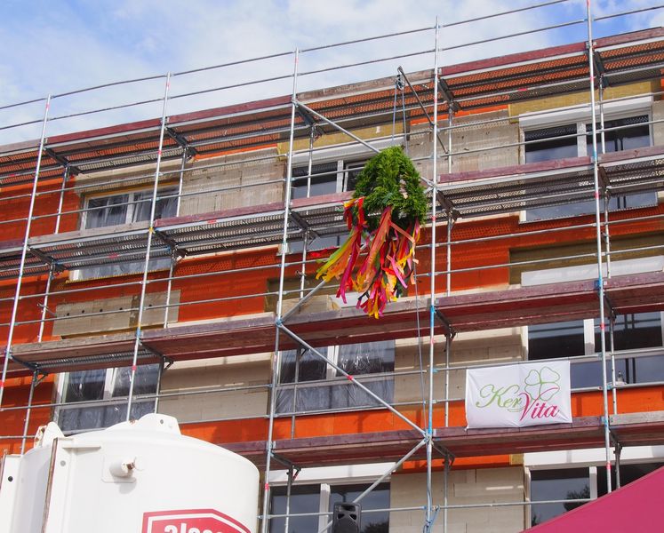 Richtfest an einem Neubau: Das Gebäude ist eingerüstet, und ein Richtkranz mit bunten Bändern hängt in der Mitte der Fassade. Ein Bauschild mit dem Logo der „KerVita Gruppe“ ist am Gerüst befestigt – die Szene symbolisiert den Baufortschritt eines Pflege-