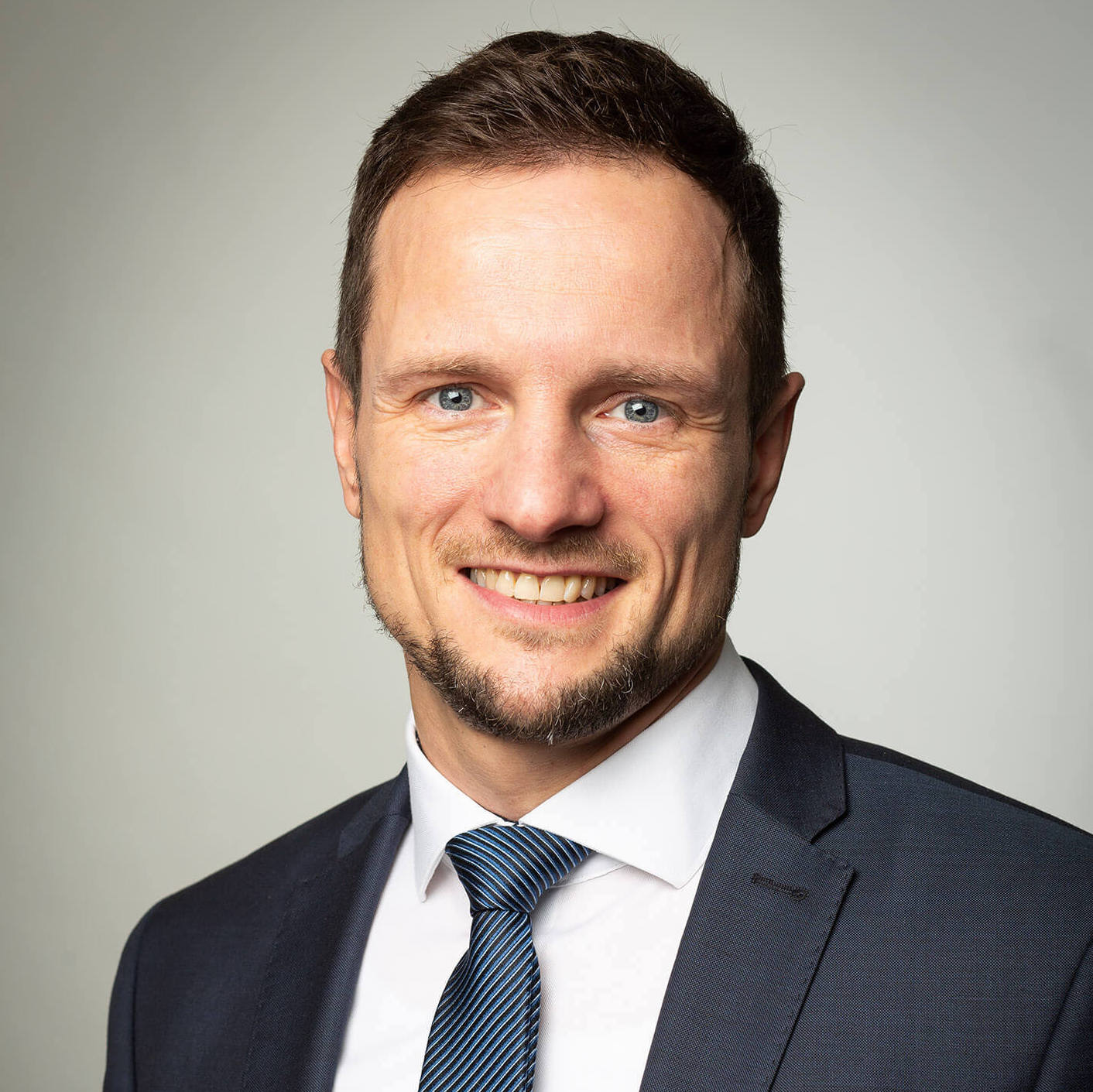 „Porträt eines freundlich lächelnden Mannes im Anzug und Krawatte vor neutralem Hintergrund – ideal für die Darstellung eines Teammitglieds oder Geschäftsführers.“