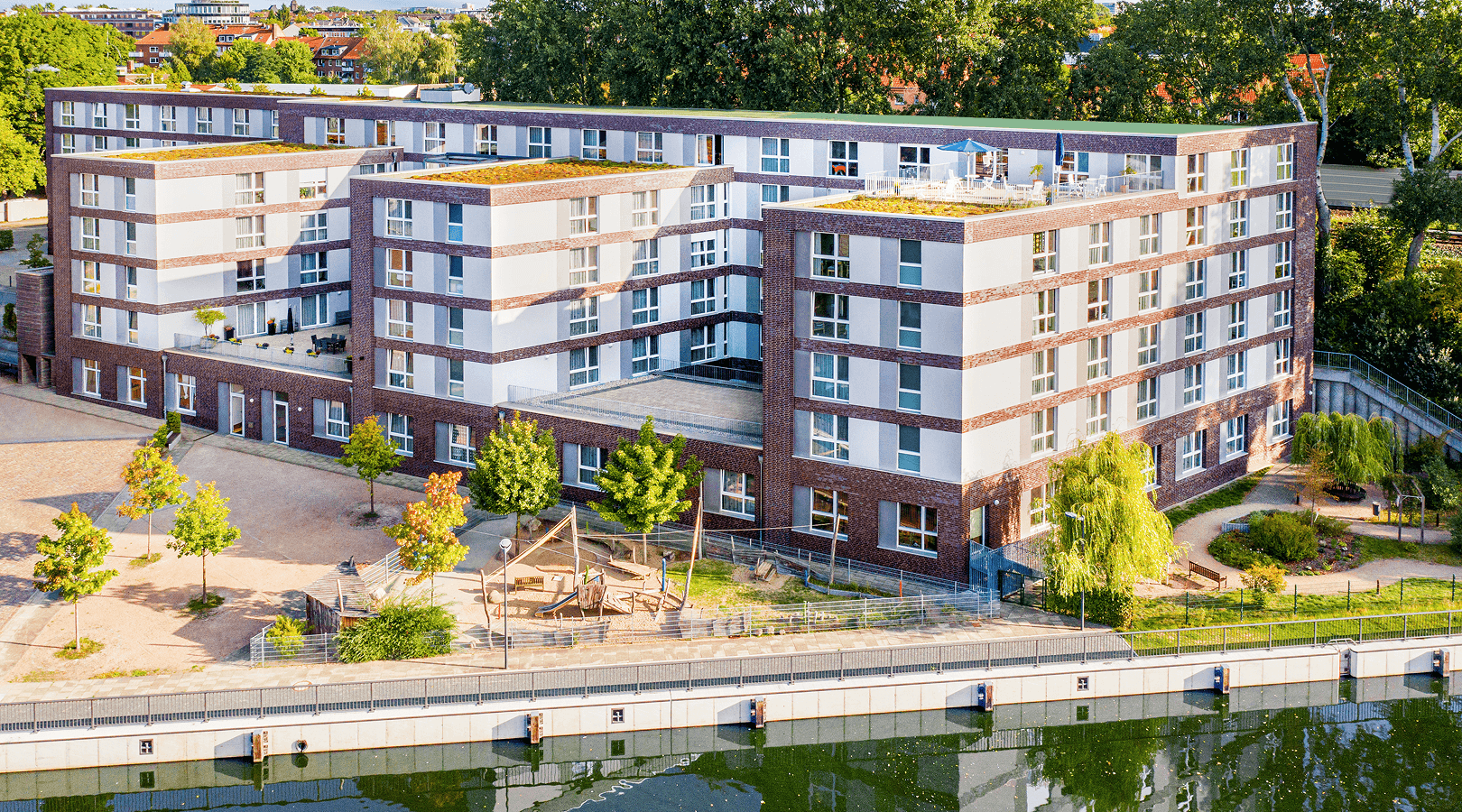 Moderne Seniorenresidenz mit Klinkerfassade und Flachdach direkt am Wasser, umgeben von grünen Bäumen und einem gepflegten Außenbereich mit Sitzgelegenheiten und Spazierwegen.