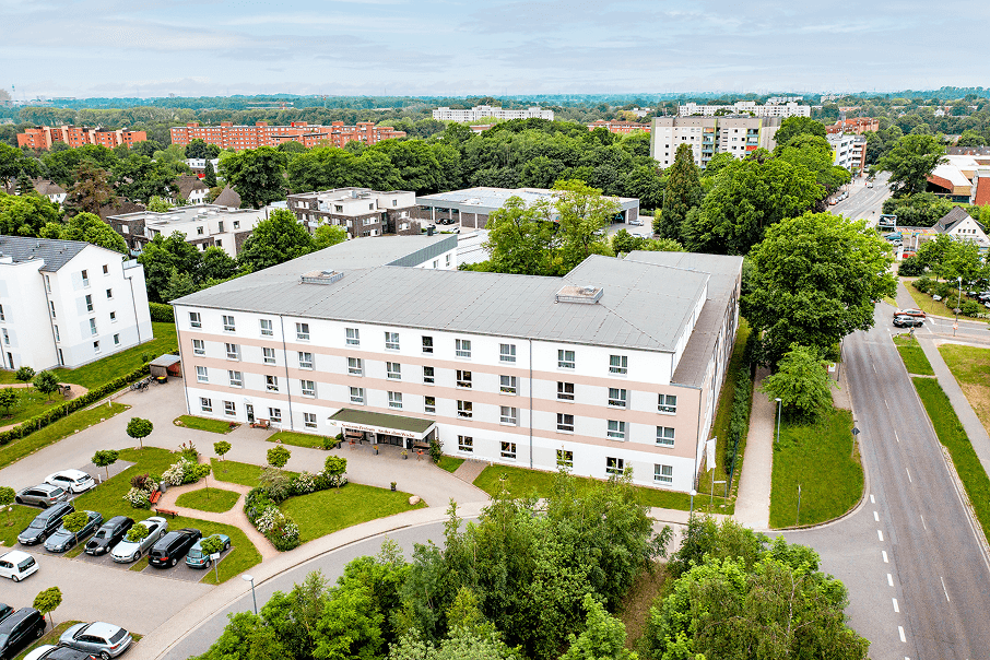 Moderne Pflegeeinrichtung in einem ruhigen Wohngebiet, umgeben von Bäumen und Grünflächen, mit großzügigem Eingangsbereich und angrenzendem Parkplatz.