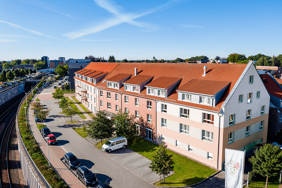 Luftaufnahme eines Seniorenheims mit hellrosa und weißen Fassaden sowie einem roten Satteldach, gelegen an einer ruhigen Straße mit angrenzendem Bahnverlauf und umliegender Wohnbebauung.