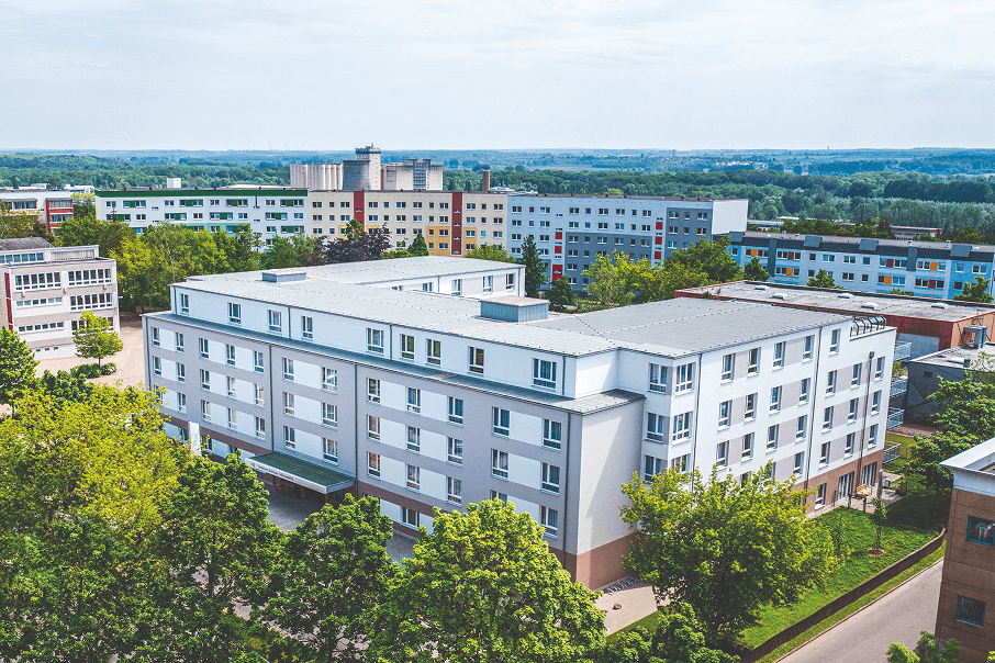 Luftaufnahme eines modernen Seniorenwohnheims mit heller Fassade, umgeben von Bäumen und Wohngebäuden in einer ruhigen städtischen Umgebung. Im Hintergrund erstreckt sich eine weitläufige, grüne Landschaft unter einem leicht bewölkten Himmel.