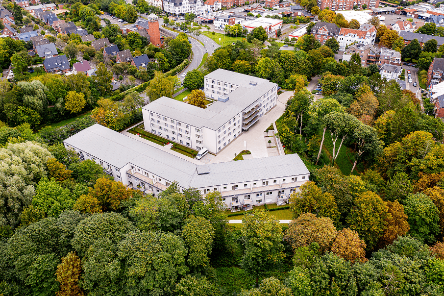Luftaufnahme eines modernen Pflegeheims inmitten einer dicht bewaldeten Umgebung mit herbstlich gefärbtem Laub. Das Gebäude hat eine helle Fassade und ist von einem geschwungenen Weg sowie benachbarten Wohnhäusern eingerahmt.